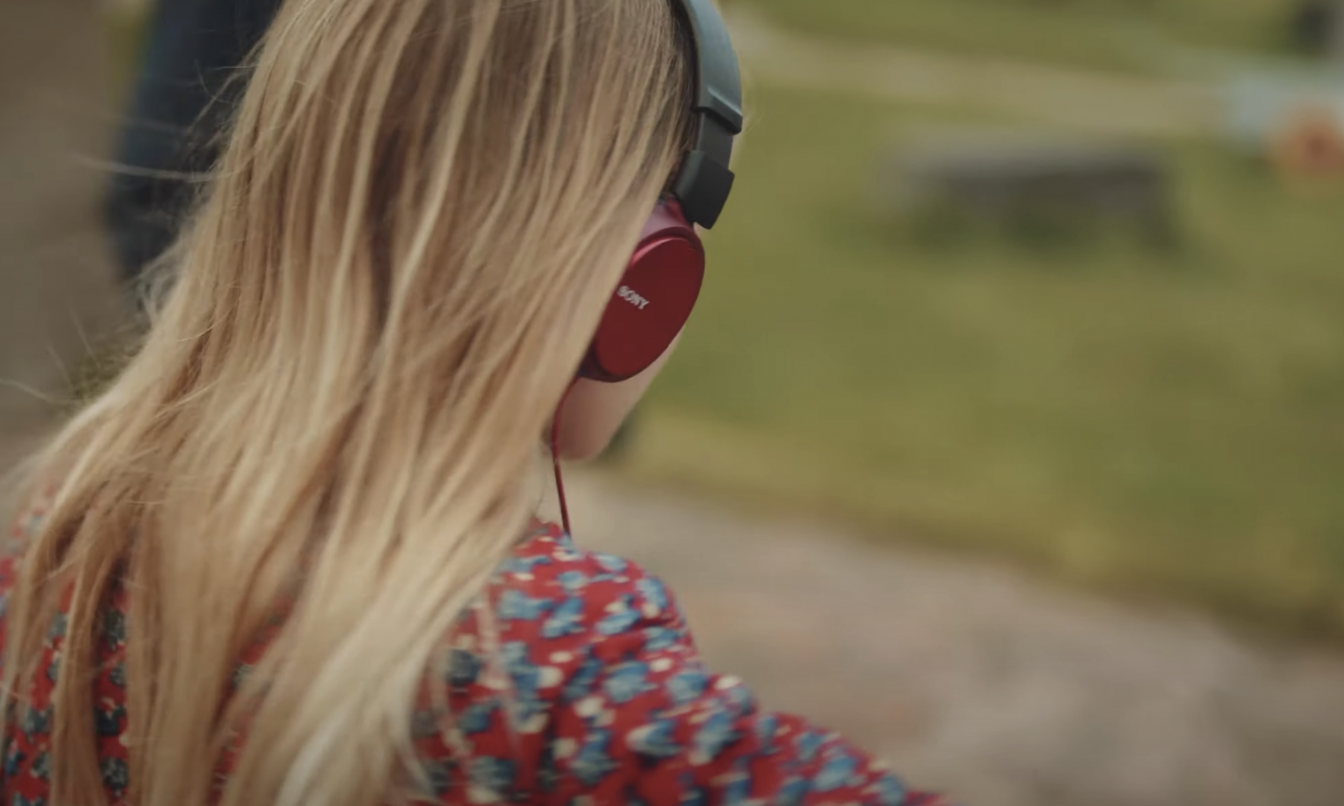 En lyshåret pige sidder med ryggen til. Hun har et par røde høretelefoner på. Hun har en rød bluse med blå blomster på. Hun kigger ud over en græsplæne med en træstub på.
