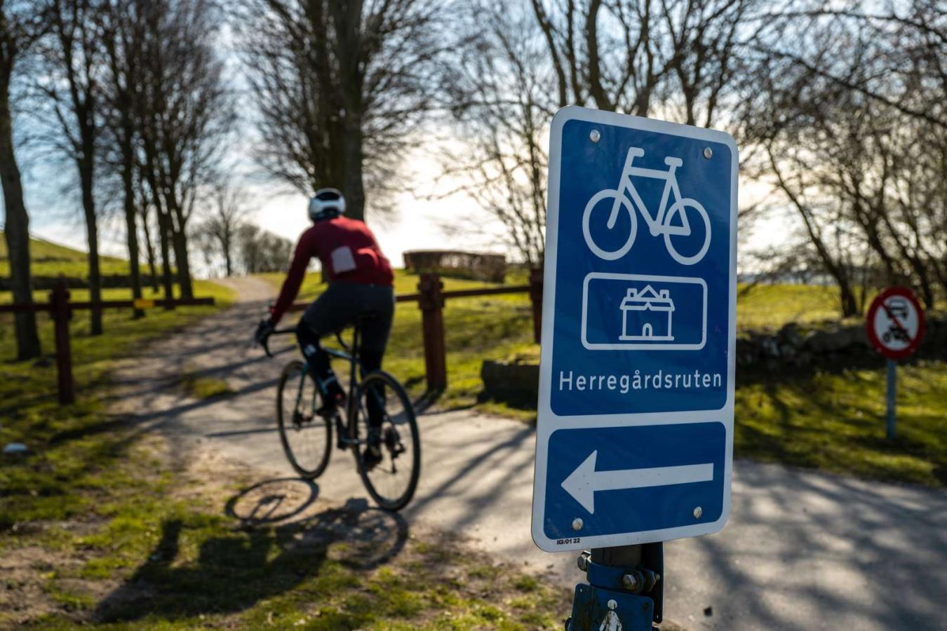 i forgrunden er et blåt skilt med en cykel, pil til venstre og tekst hvor der står Herregårdsruten. I baggrunden cykler en mand på racercykel ned af en sti der går mellem træer. 