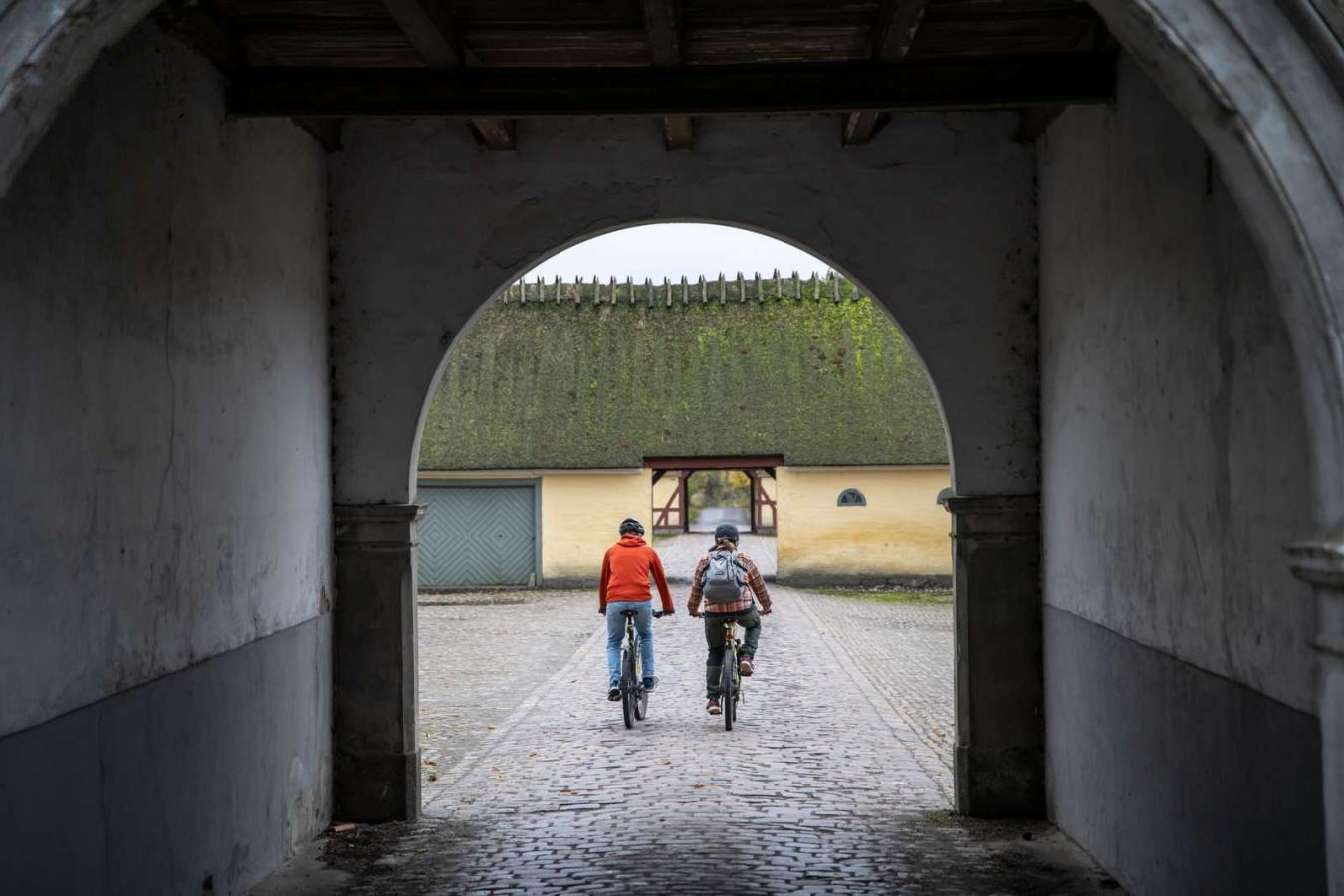 To cyklister ses gennem buet port på brostensbelagt gårdsplads.