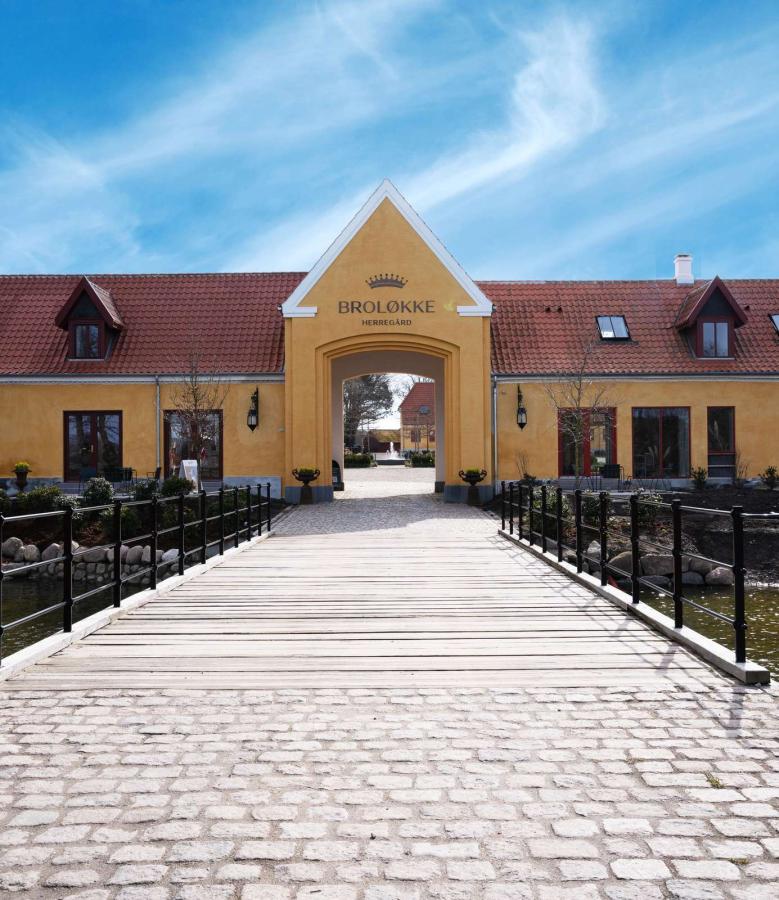 Porten ind til Broløkke Herregård. Gul bygning med rødt tag og træbro hen mod porten