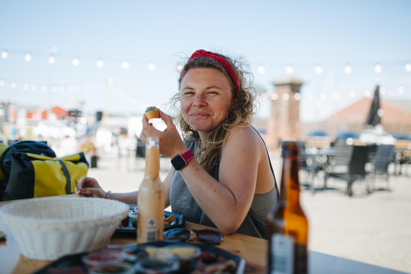 Kvinde med snack i hånden sidder med og mad og drikke på bordet foran og smiler til kameraet.