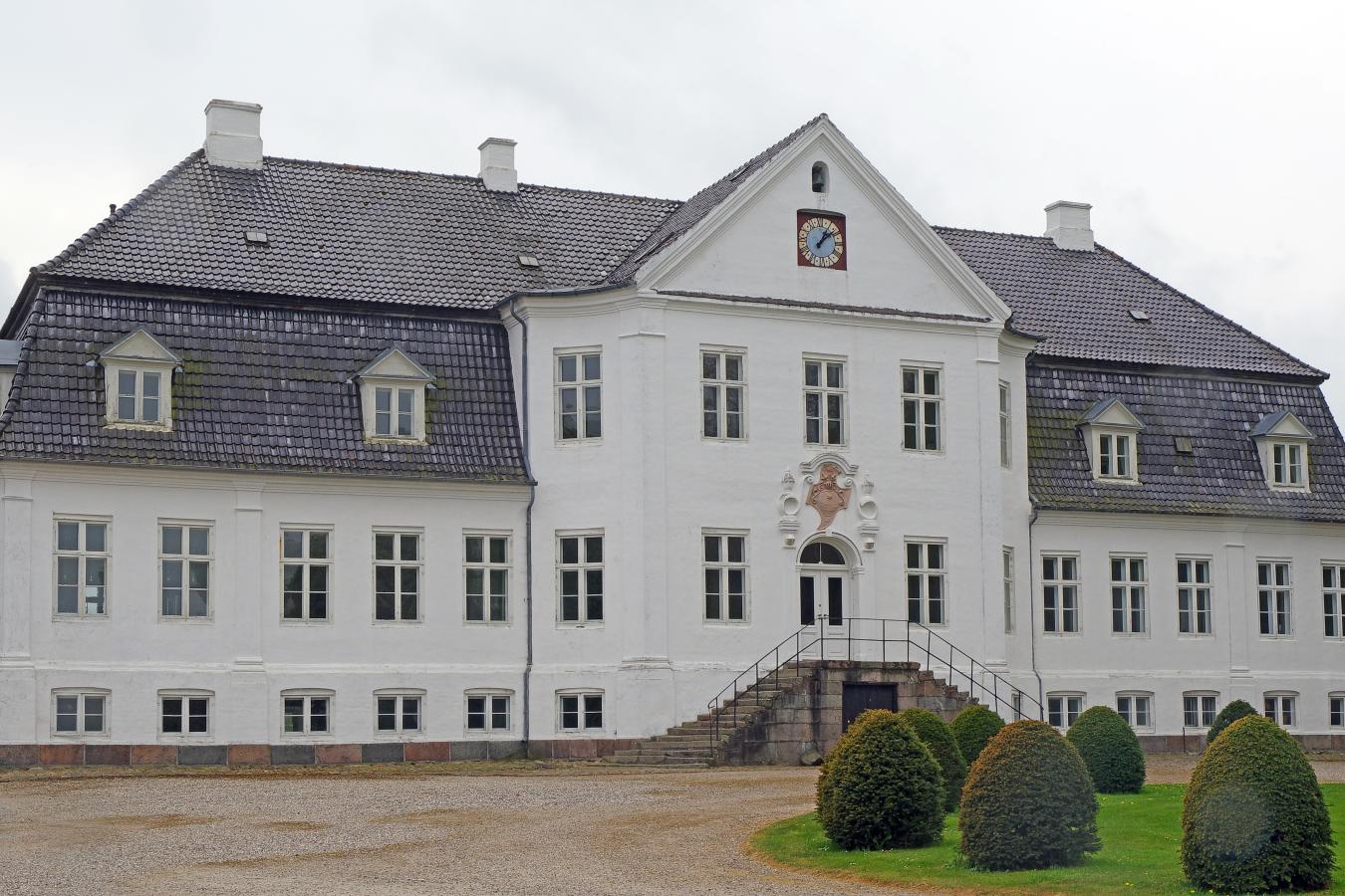 Stort hvidt gods står midt i billedet. Taget er sort og over hovedindgangen hænger en farvet figur. En bred trappe fører op til hovedindgangen. Foran trappen er kanten af en græsplæne, hvor der står 7 runde buske.