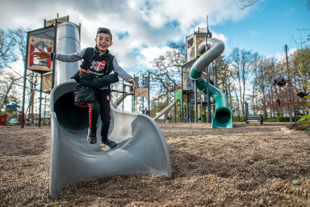 Smilende dreng står på rutsjebane med en legeplads i baggrunden.