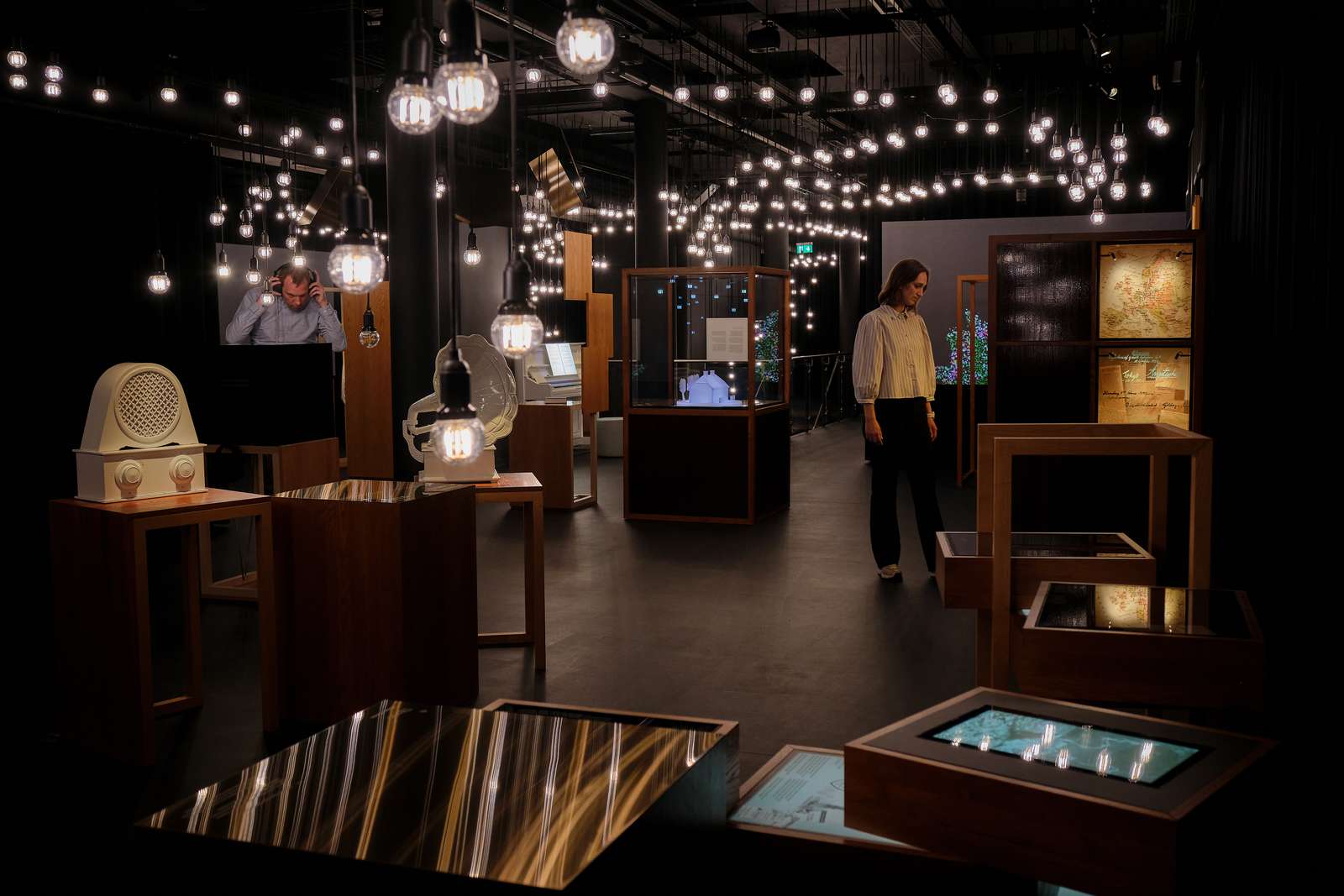 En kvinde og en mand udforsker en udstilling. Lokalet er mørkt med udstillingsmontrer og en masse lamper med lysende pærer i loftet.