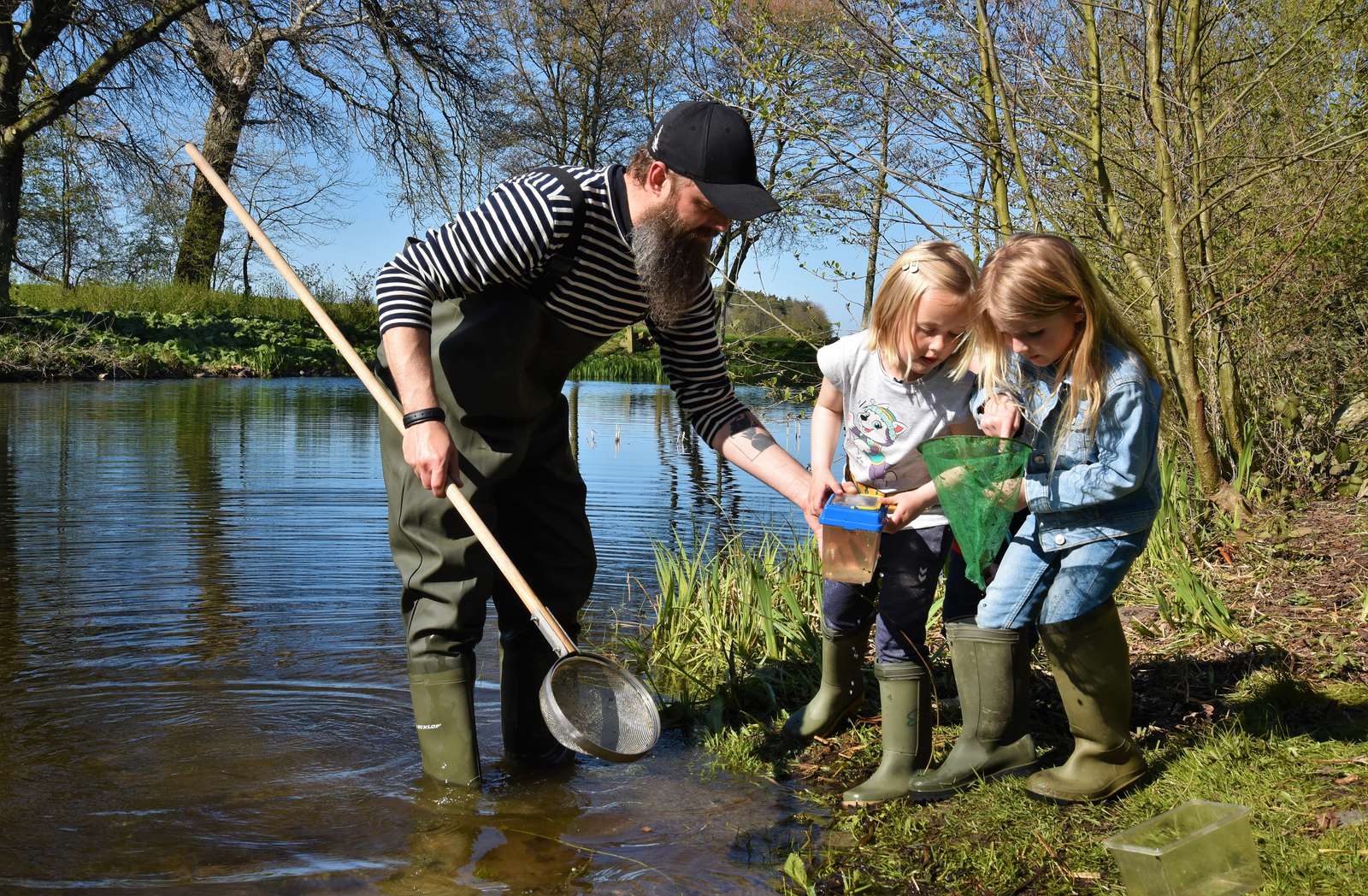 To børn og en voksen står ved breden af en å og kigger på, hvad de har fået i nettet.