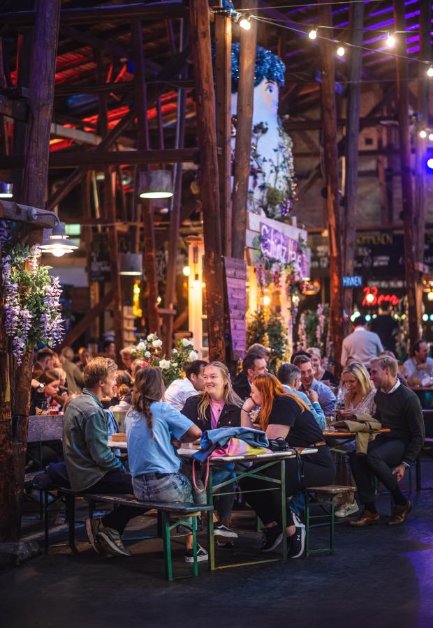 En gruppe mennesker sidder i et pakhus og hygger og spiser mad.