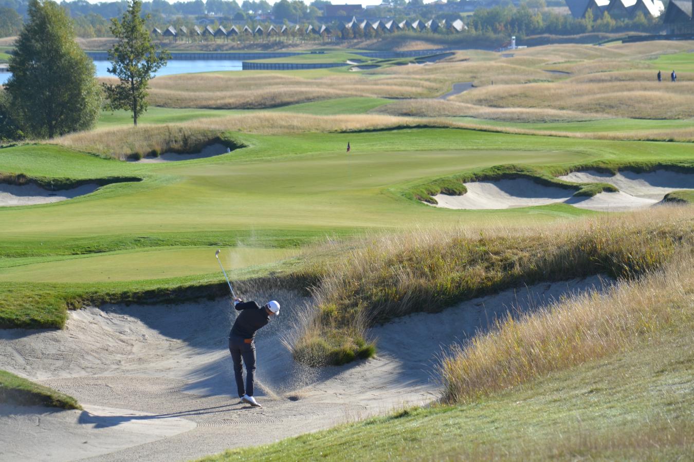 Golfspiller på bølgende grøn golfbane slår et slag fra en bunker, så sandet står op.