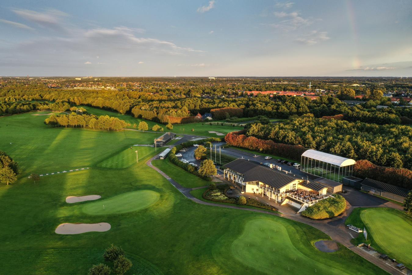 Udsigt over grøn golfbane oppefra med golfklubbens lokaler i forgrunden og Odense by i baggerunden.