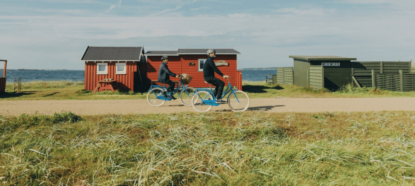 Et midaldrende par cykler på en grusvej foran et par farverige badehuse med havet i baggrunden