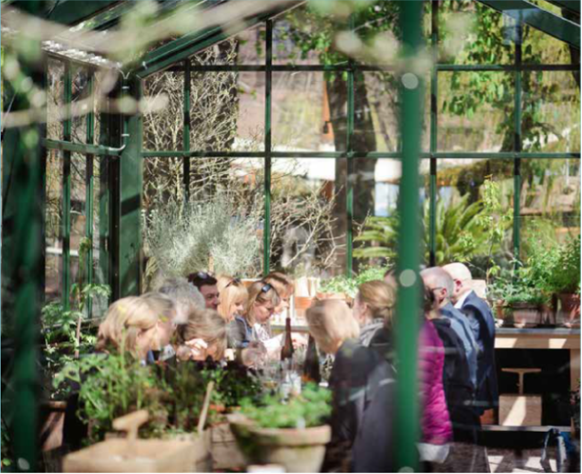 En flok mennesker sider ved et langt bord i en drivhuslignende bygning i grønne omgivelser.