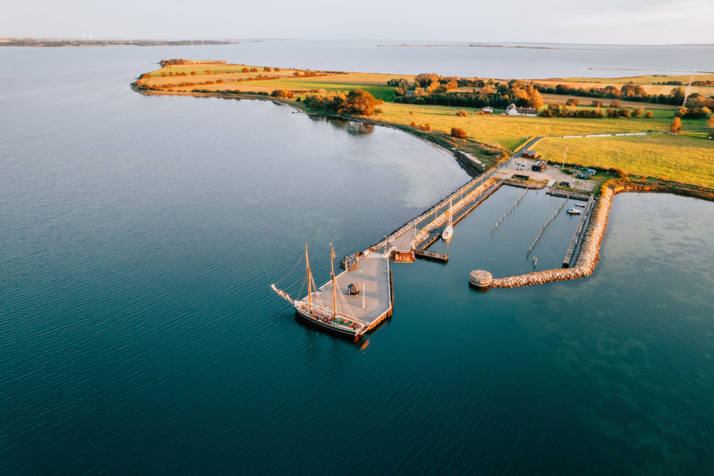 Dronefoto af Skarø havn, hvor en tomastet skonnert ligger som den eneste båd for enden af havnen