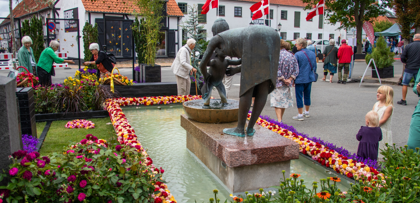 Billede af springvand med statue i midten, dekoreret med kant af roser i gule, orange og røde farver.