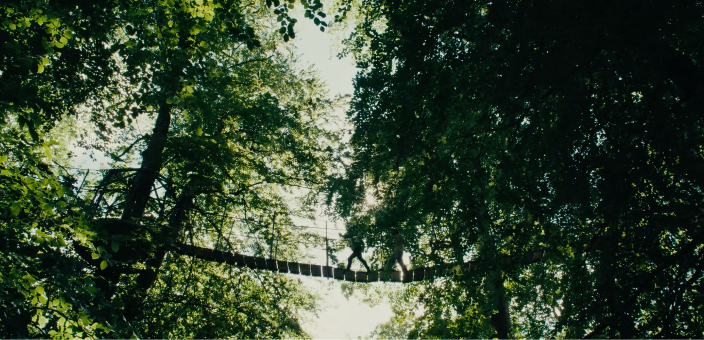 Mellem høje grønne trækroner hænger en hængebro, hvor to personer er på vej over