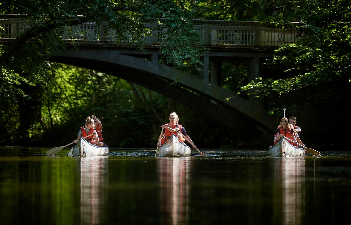 Tre kanoer med to personer i hver roer på en å omkranset af grønne træer og med en bro i baggrunden