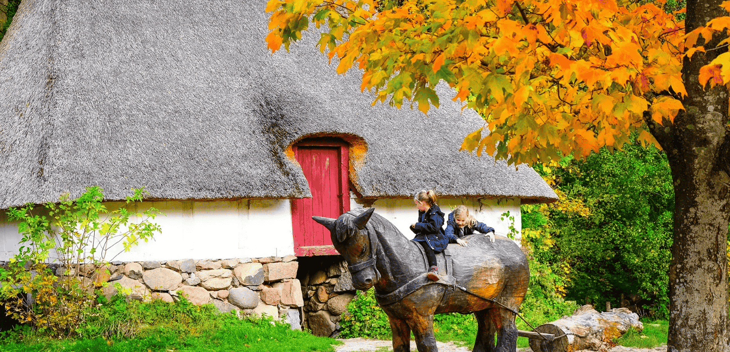 Børn leger på en hest udskåret af træ foran et gammelt stråtækt hus og et træ med gul-orange blade på
