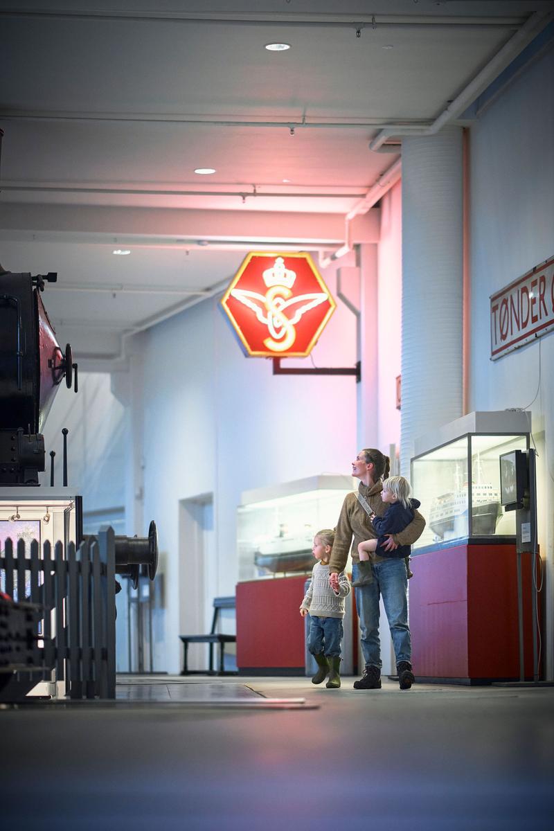 En mor holder sit barn i armen og holder i hånden med det andet barn, mens de går rundt på en udstilling på jernbanemuseet