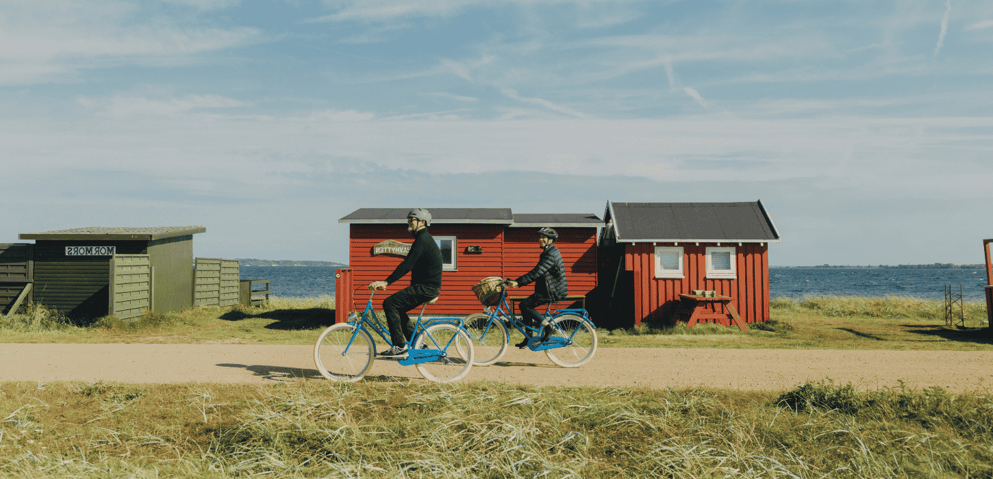 To cyklister kører på en grusvej med græs til den ene side og hav og badehuse til den anden side