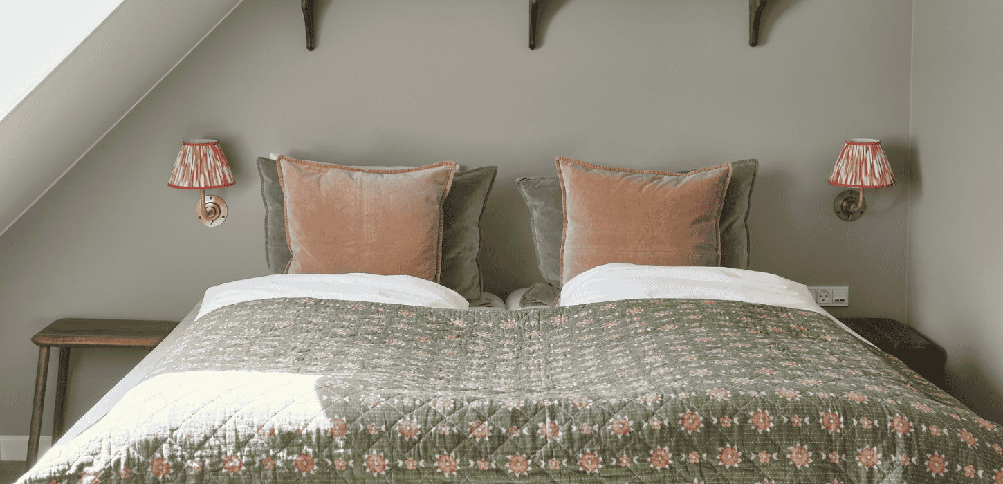 En opredt dobbeltseng med lyserøde pyntepuder i et smalt værelse med skråvæg
