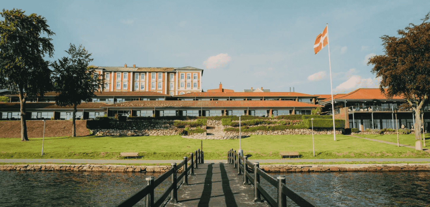 Et rødmuret hotel der ligger på et grønt område lige ned til vandkanten, taget fra en badebro