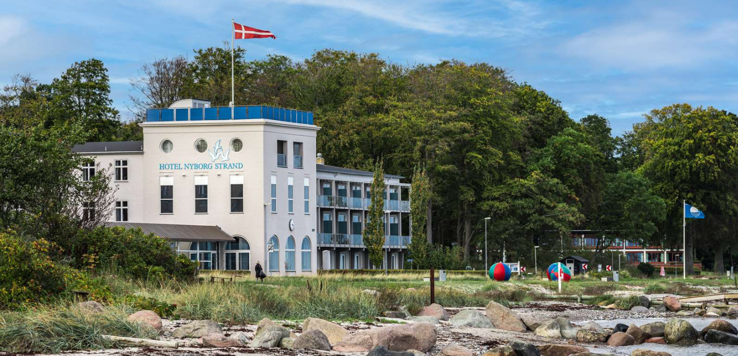 En hvid bygning beliggende tæt ved strandkanten med skov og grønne træer i baggrunden