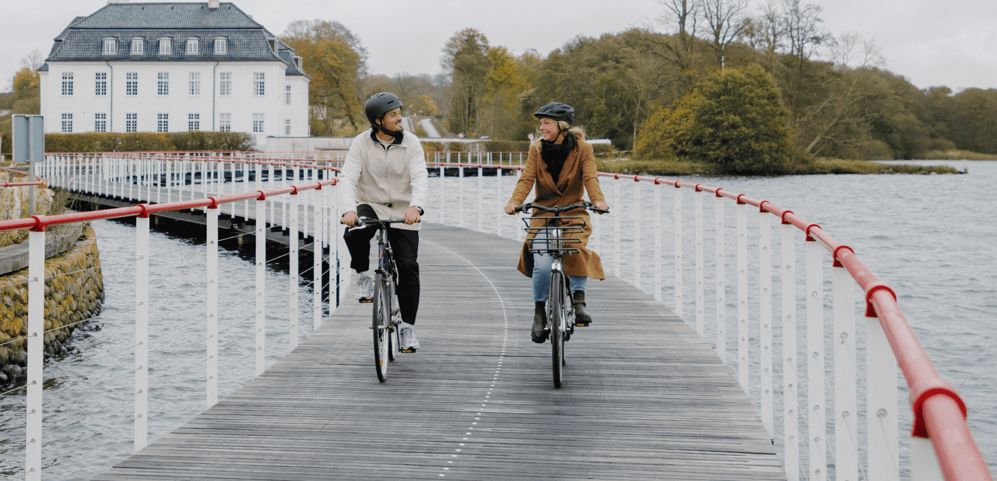 Two cyclists are riding on a bike path built on water, with a white manor house in the background
