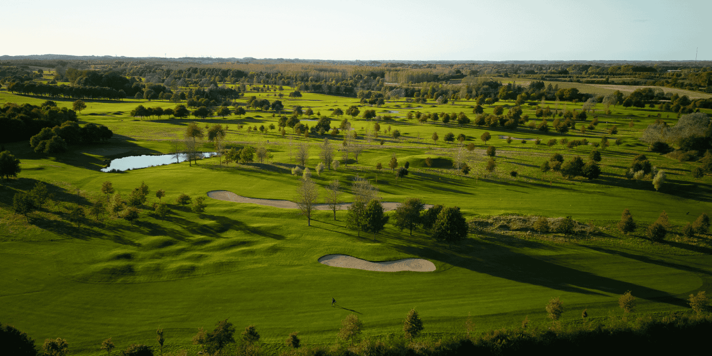 Grøn golfbane med grupper af træer, bunkers og en lille sø