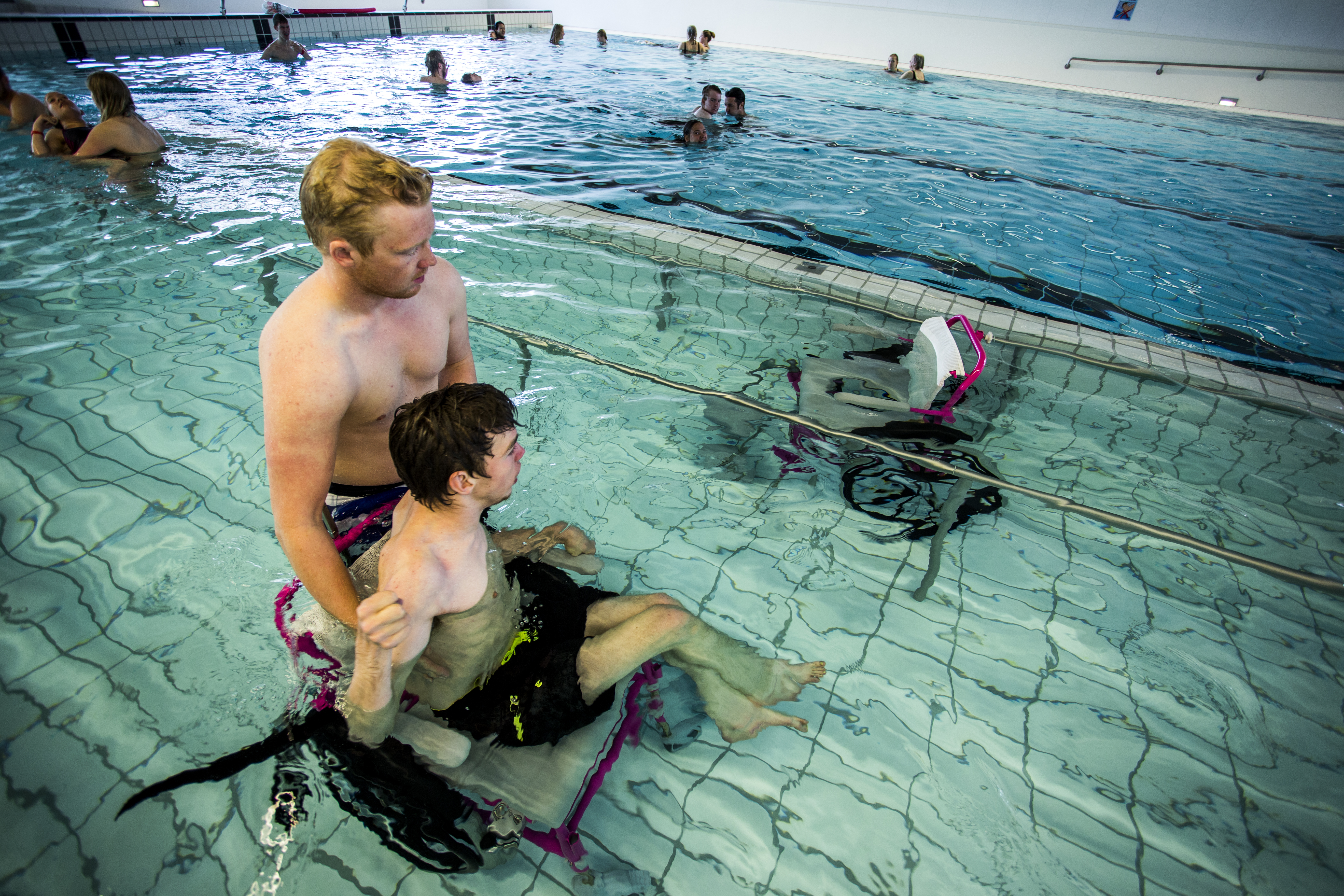 To mænd i en svømmehal hvor den ene er i kørestol i vandet