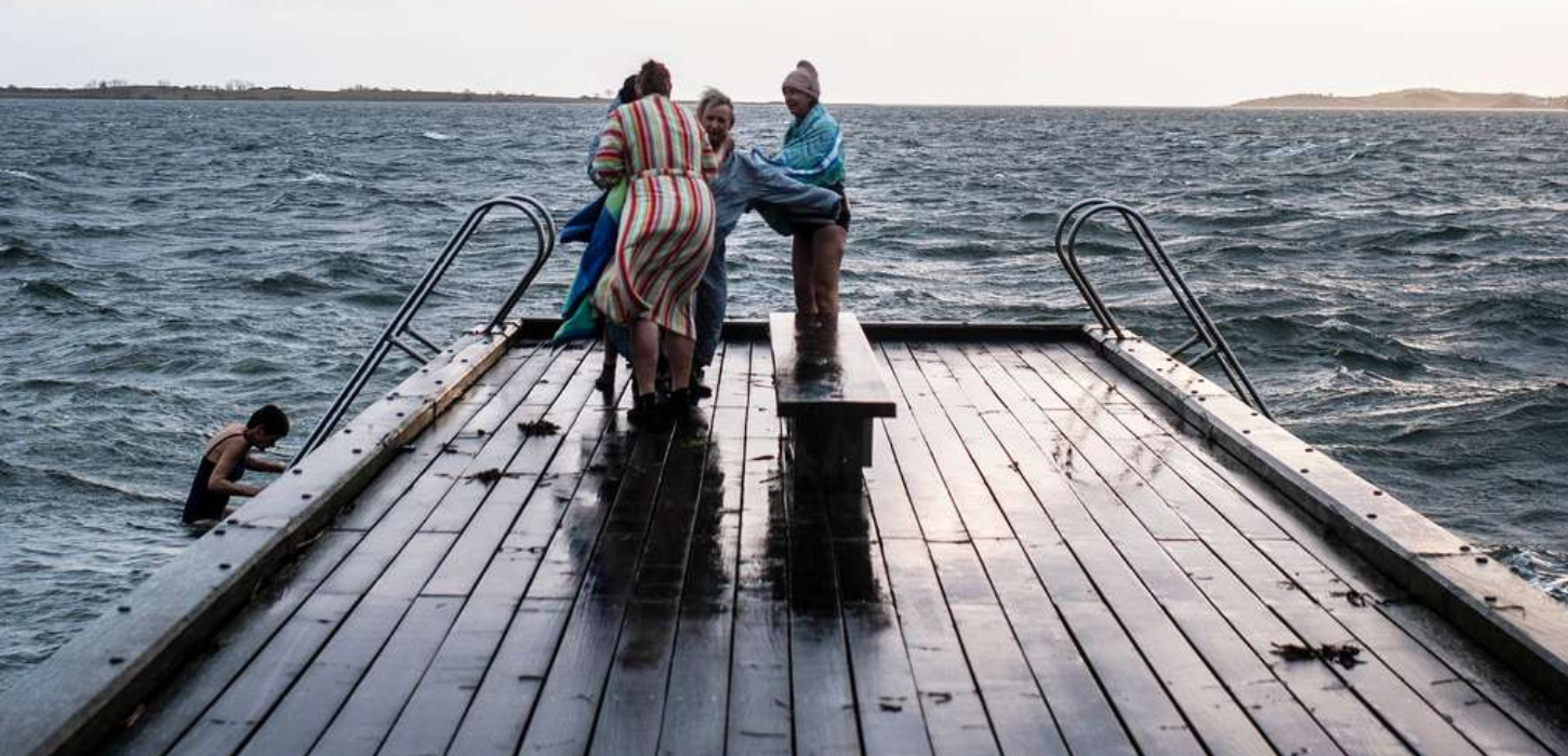 En gruppe vinterbadere står på en bred badebro i skumringen omkranset af et bølgende hav