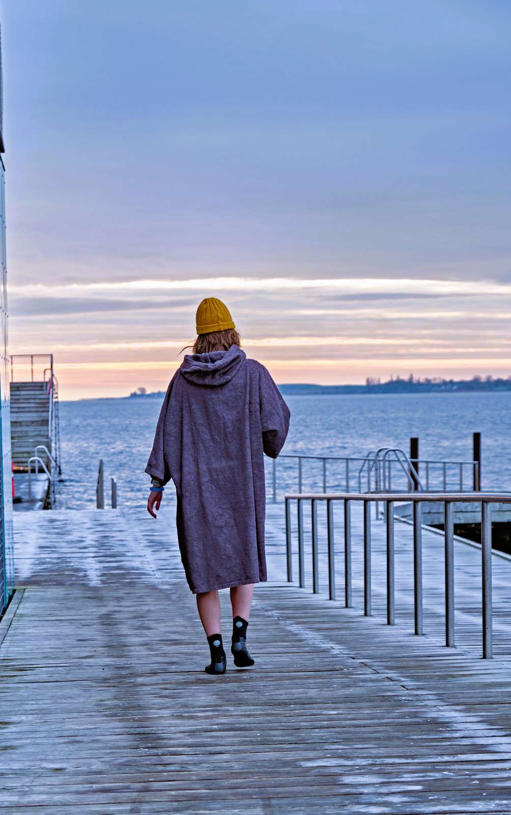Kvinde iført vinterbadermorgenkåbe går ud af en rimbelagt badebro i solnedgangen