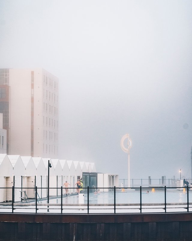 Et havnebad i en tæt tåge med hegn rundt om og hvide små huse ved siden af bassinet