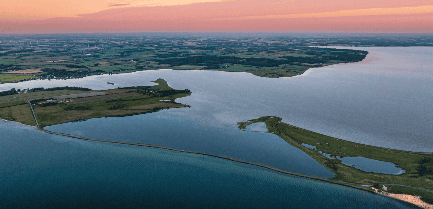 Et luttfoto af en dæmning med havvand på begge sider i solnedgang med lyserød himmel 
