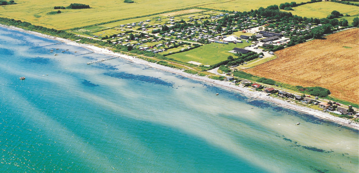 Luftfoto af Camp Hverringe, som ligger omkranset af marker lige ud til en hvid og lavvandet strand