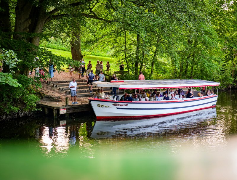 En hvid båd lægger til en lille afsats i en grøn skov, mens et par mennesker 