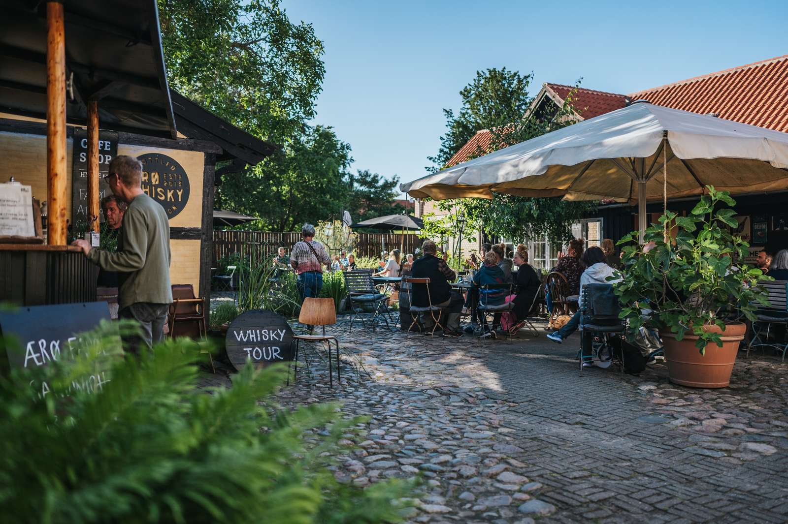 En brostensbelagt gårdhave med servering, parasol og store plantekrukker