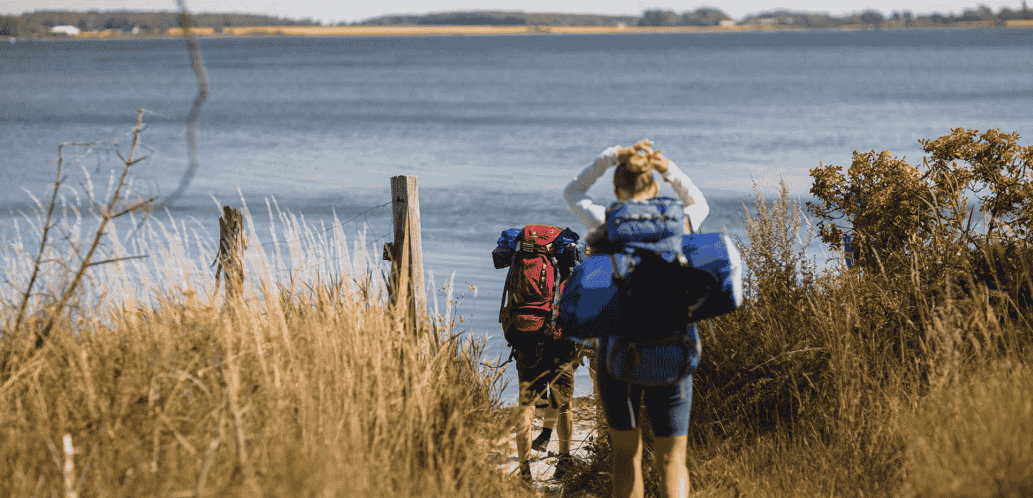 To vandrere er på vej igennem højt græs ned til havet, og de bærer begge vandreudstyr og rygsæk