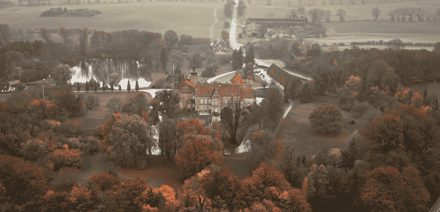 Dronefoto af et slot med spir og tårne placeret midt i et efterårsfarvet landskab i grønne og orange farver