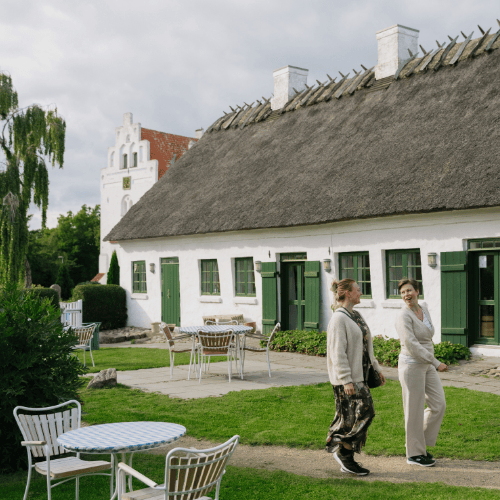 To kvinder går foran en gammel kirkelade med stråtækt tag og en hvid kirke i baggrunden