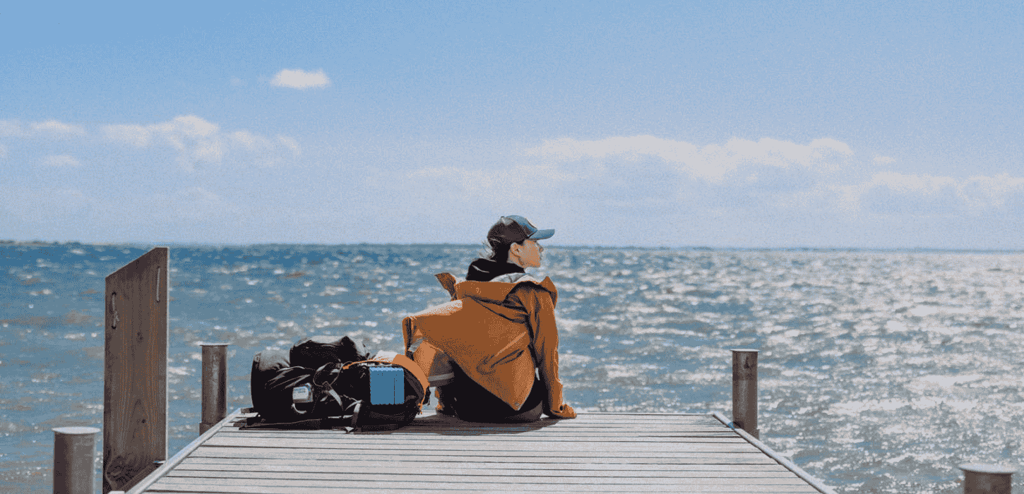 En kvindelig vandrer sidder iført regntøj på badebro med udsigt til himmel og hav