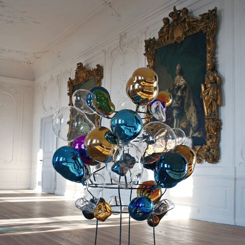 En kunstinstallation af en masse deforme glaskugler i forskellige farver, placeret i en sal med gamle guldindrammede portrætter