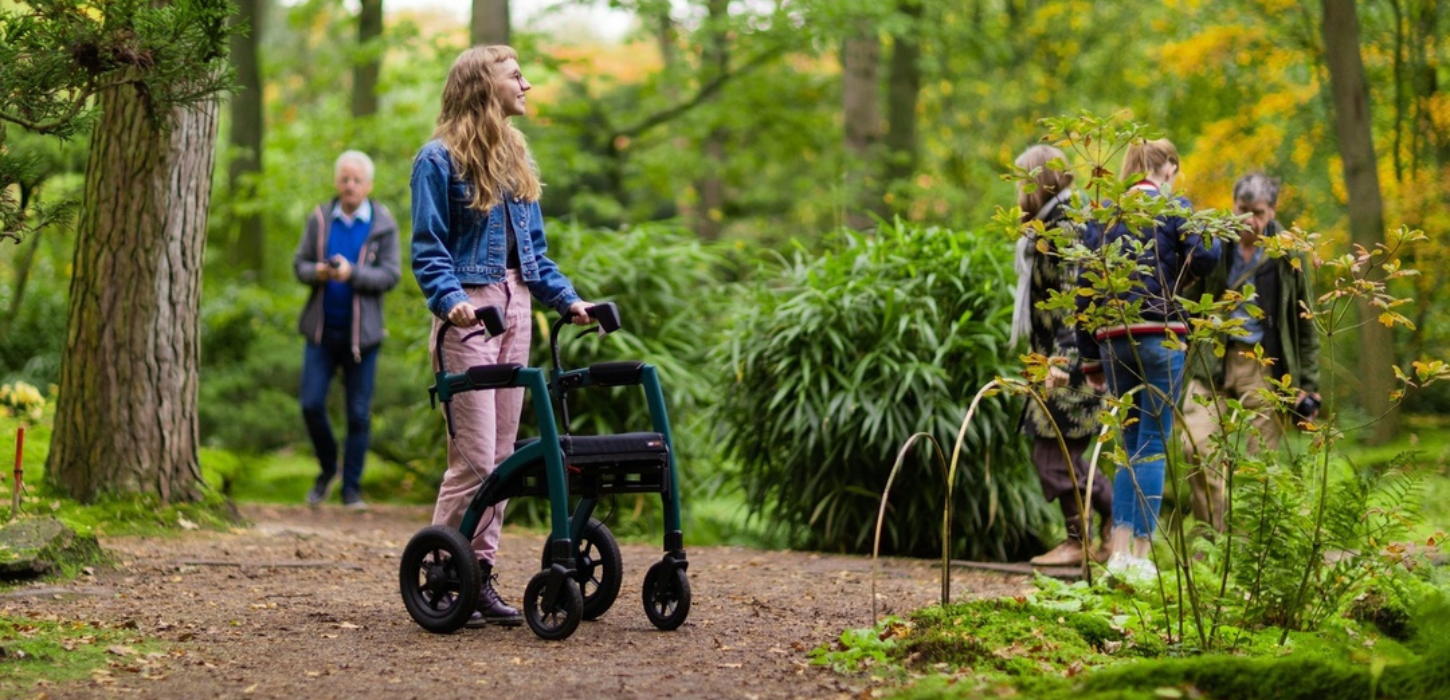 Ung pige med rollator står i grønne omgivelser med andre mennesker i baggrunden
