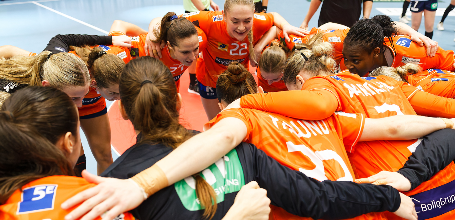 Håndboldkvinder i orange spillertøj danner rundkreds med armene om hinanden