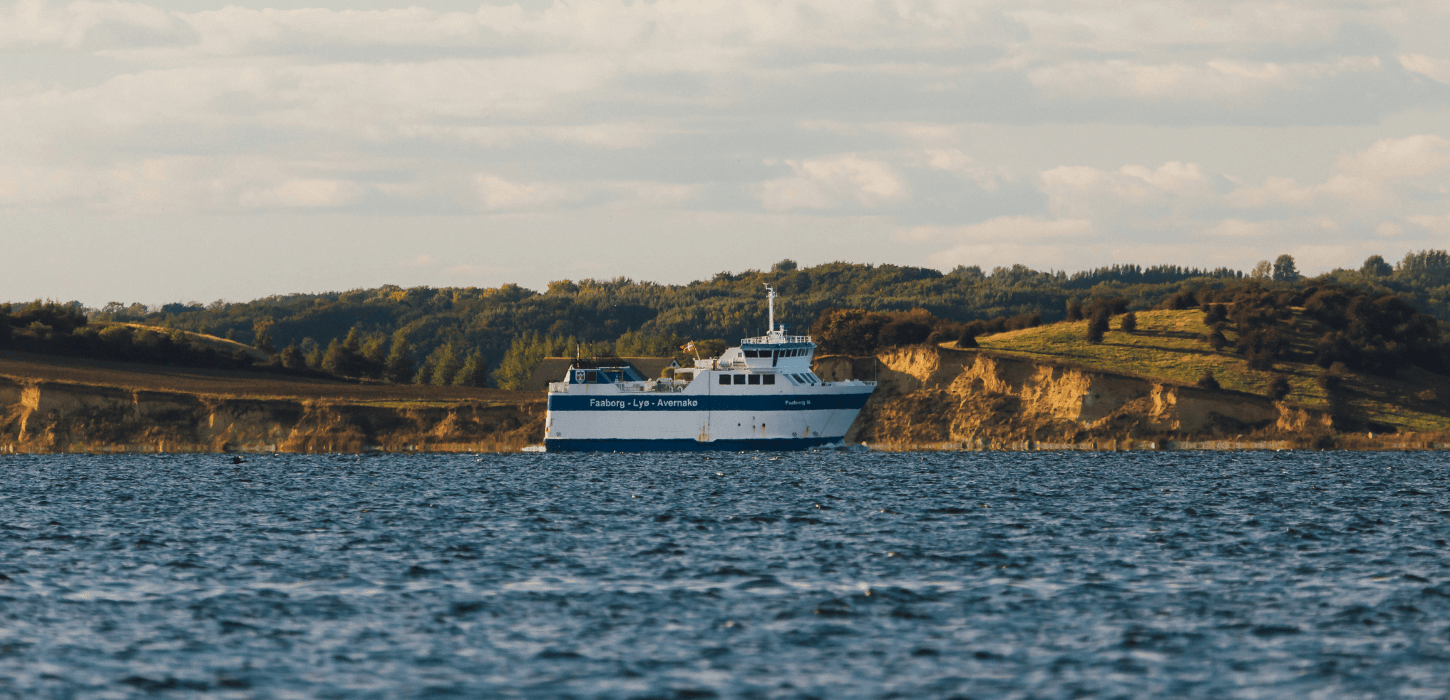 En hvid og blå færge sejler over et farvand i det maleriske Sydfynske øhav med en kuperet, grøn kystlinje i baggrunden under en overskyet himmel.