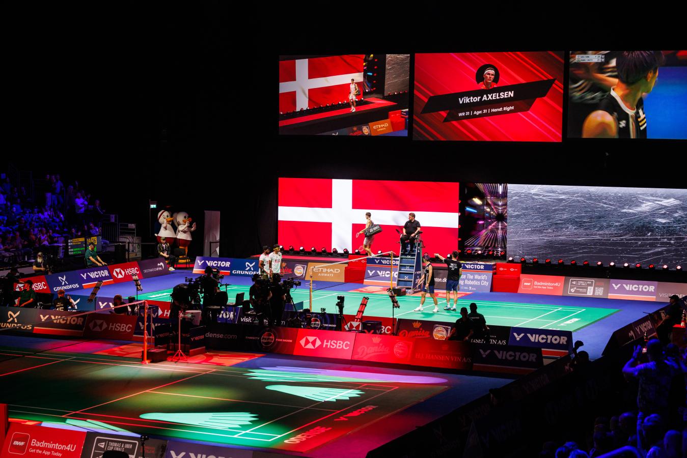 Oplyst badmintonarena med danske flag og Viktor Axelsen på vej ind på banen