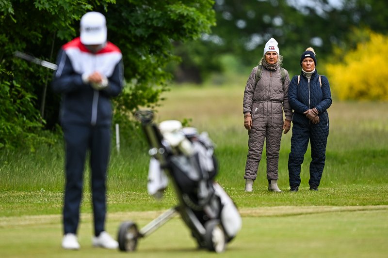 En golfspiller står med sin golftaske på en græsplæne med to tilskuere i baggrunden