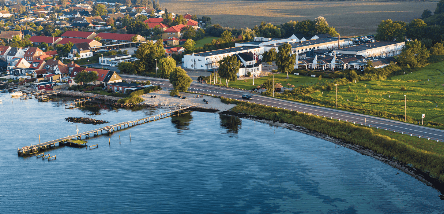 Dronefoto af hotel lige ud til kysten med grønt græs, blåt hav og et rødt svajende dannebrog