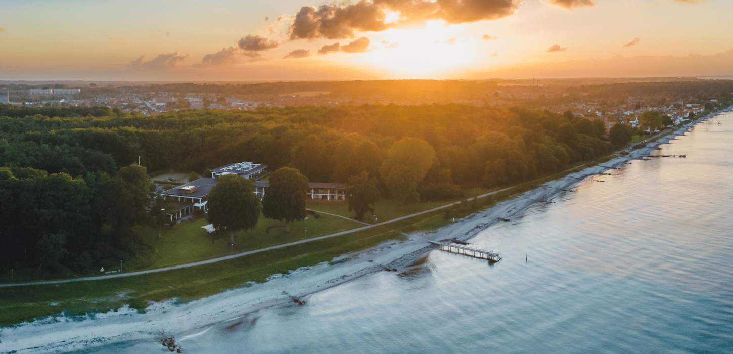 Et dronefoto af Hotel Hesselet, som ligger lige ud til kysten, set under en solnedgang med orange nuancer i horisonten bagved hotellet