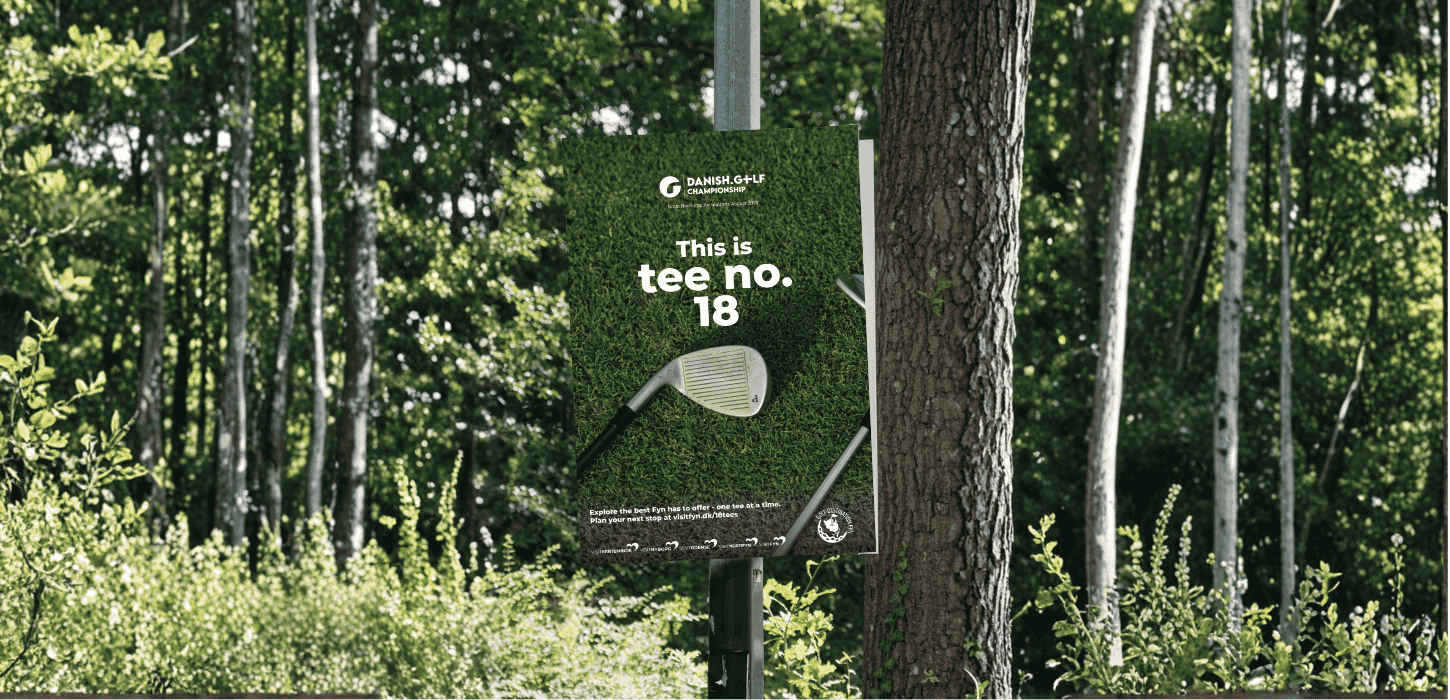 En plakat med teksten "This is tee no 18" hænger i en lygtepæl på en baggrund af træer og grønne blade