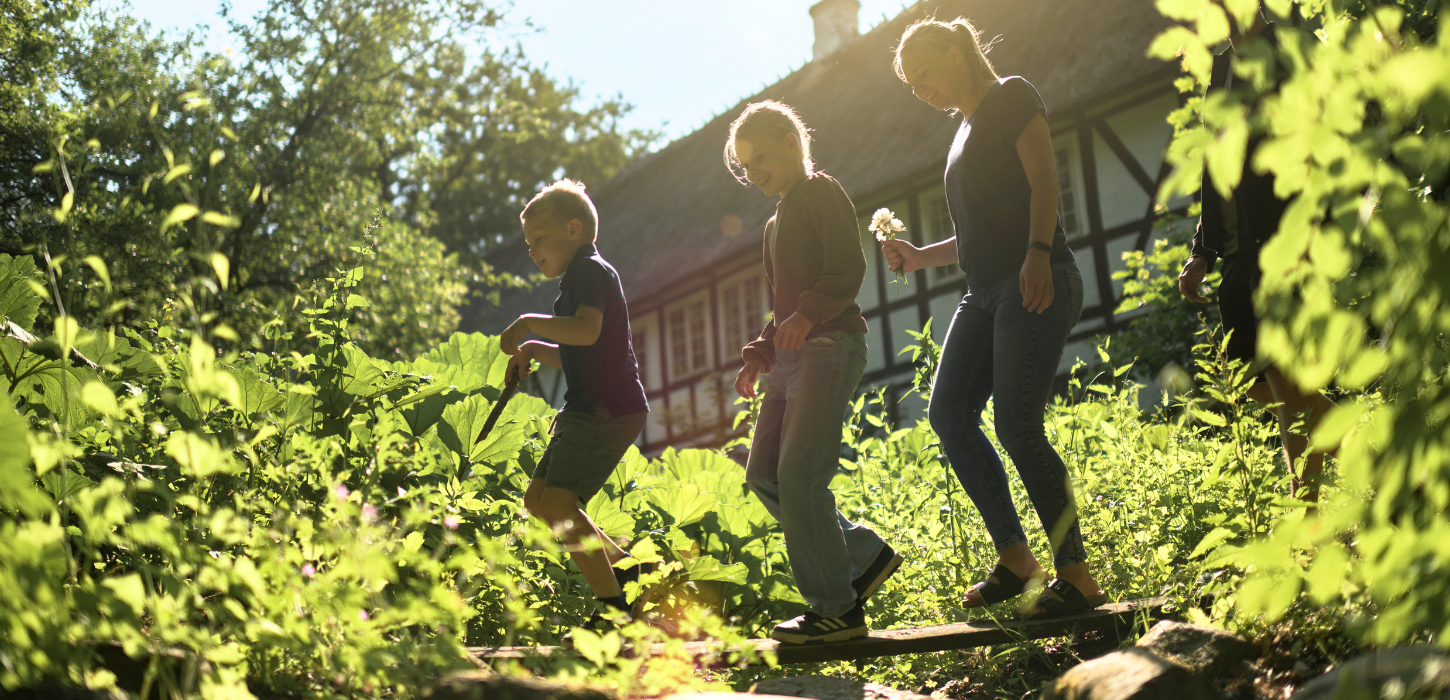 To børn og en kvinde går på et bræt midt i et grønt område med et gammeldags bindingsværkshus i baggrunden. 