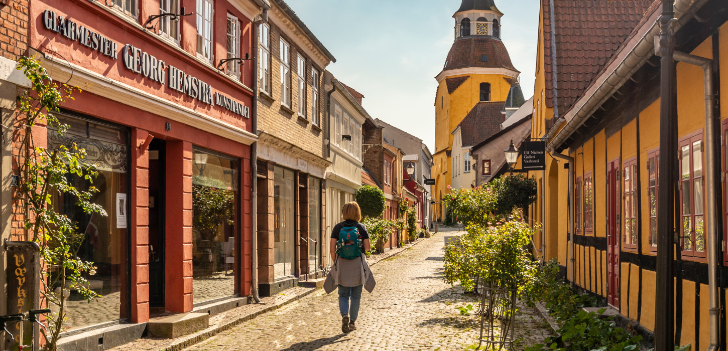 En kvinde med rygsæk går på en smal gade i en by med farverige bindingsværksfacader og et gult klokketårn i baggrunden